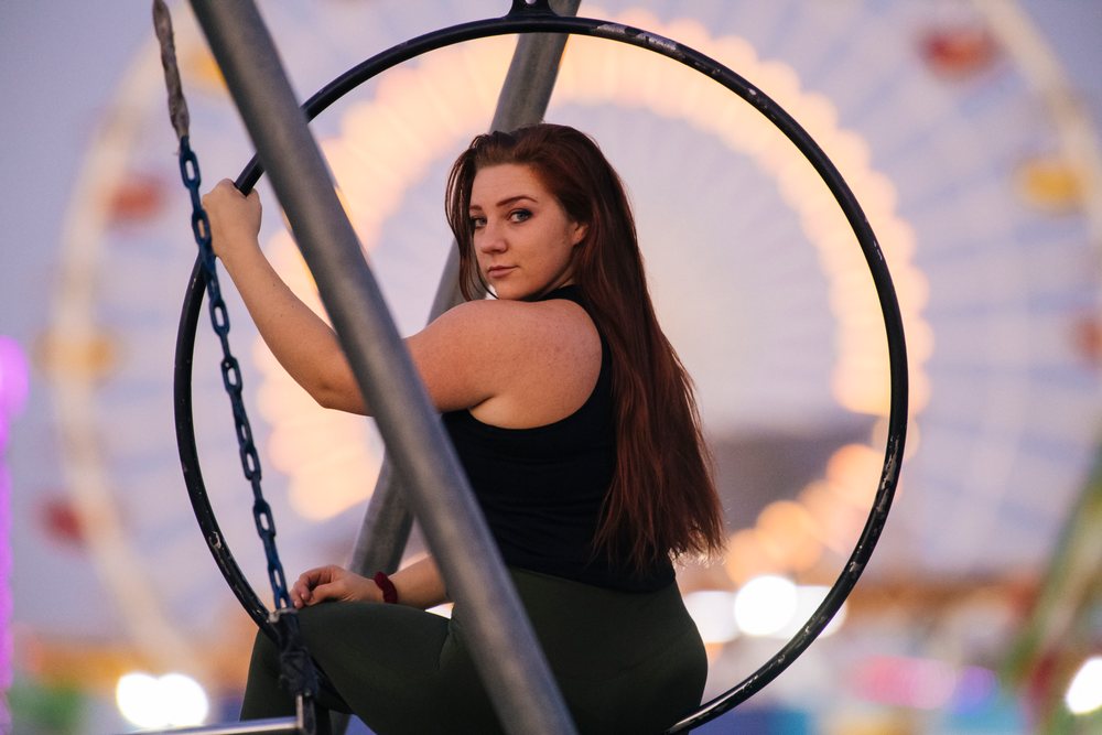 Adult woman with long hair looking back while sitting on a metal-framed swing at an amusement park with a ferris wheel in evening, California, USA.