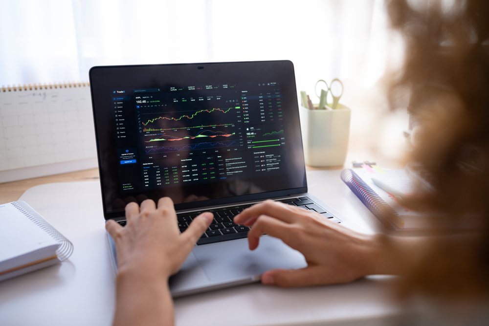 Hands typing on a laptop showing stock market charts and financial data in a home office desk workspace, Spain.