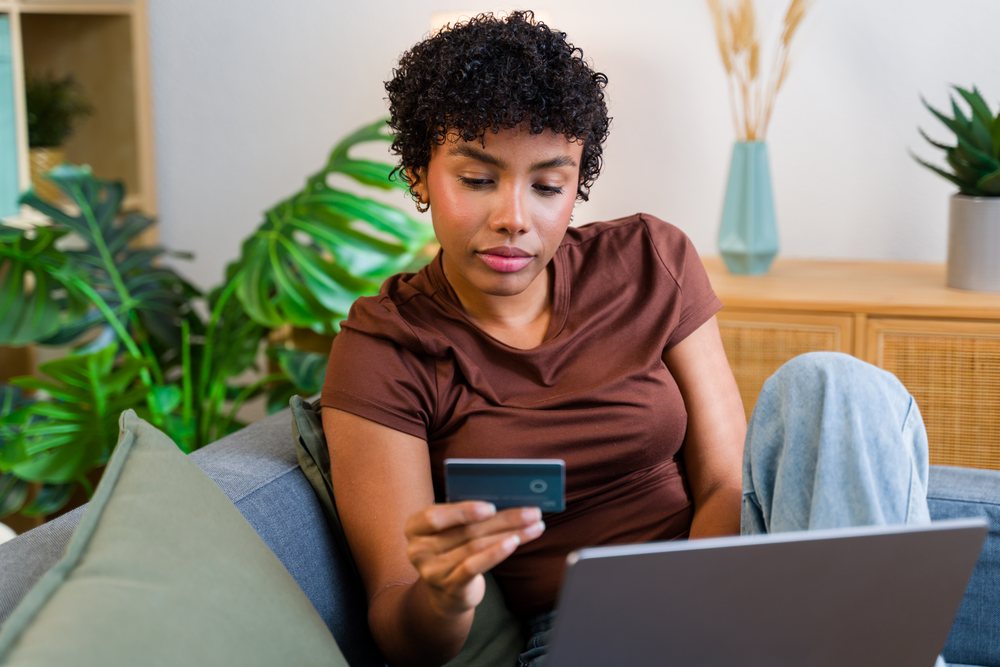 Woman sitting on a gray couch in a living room using a credit card and laptop for online shopping, Spain.