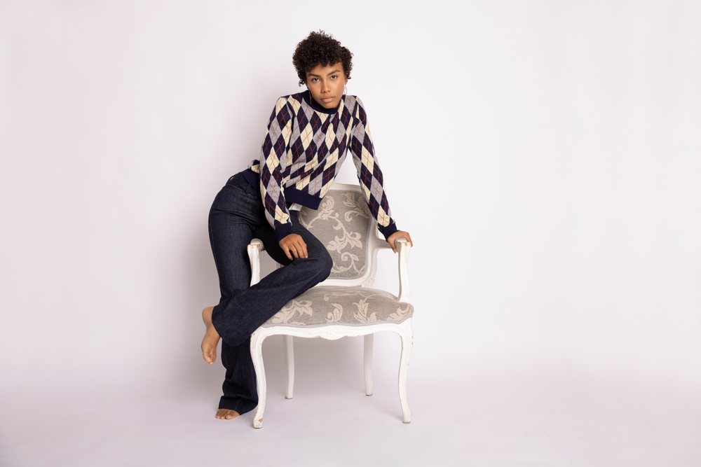 Young woman wearing an argyle sweater and dark jeans poses barefoot leaning on an armchair in a studio with a white background, fashion, young culture.