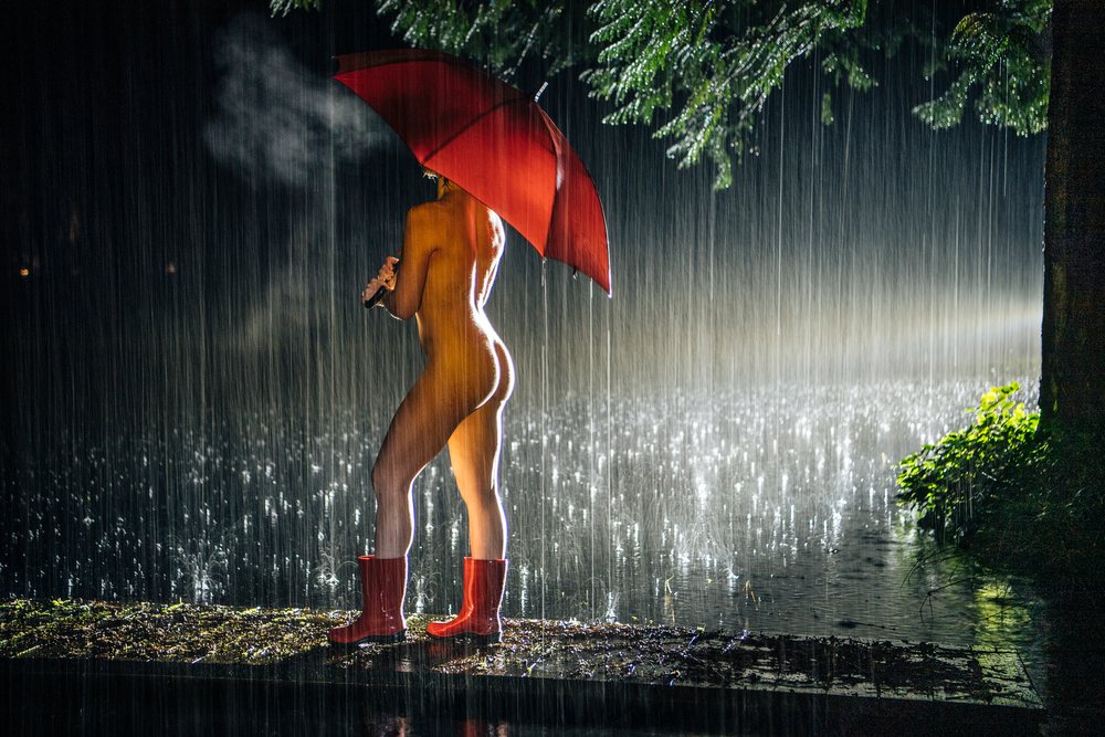 Naked woman stands holding a red umbrella and wearing red boots near a tree on wet ground in heavy rain at night, cosplay, nudity, freedom, expression, USA.