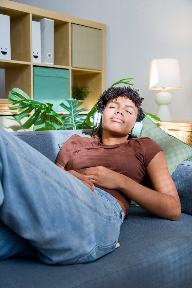 Young woman lying on a couch wearing headphones in a cozy living room with indoor plants and natural light.