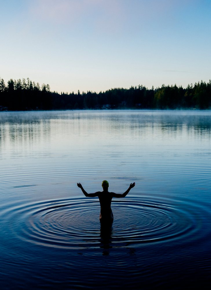 Person wearing a swim cap standing in calm lake water during early morning with a forest and mist rising in the background, USA.