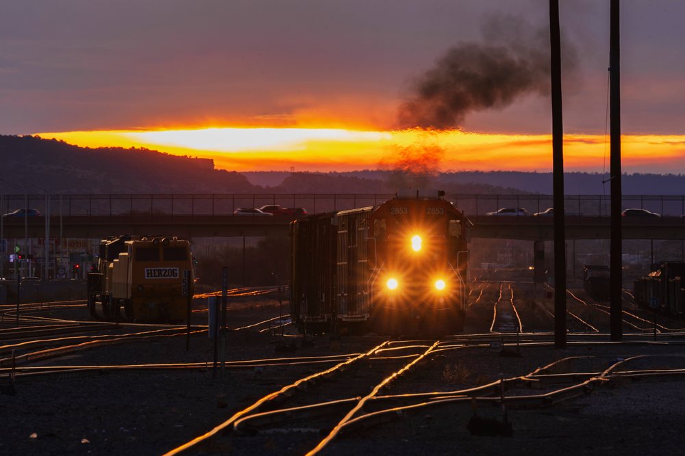 Train with freight cars and smoke moves along railway switch yard tracks under a bridge with vehicles at sunset, Americana, Historic, Gallup, New Mexico, USA.