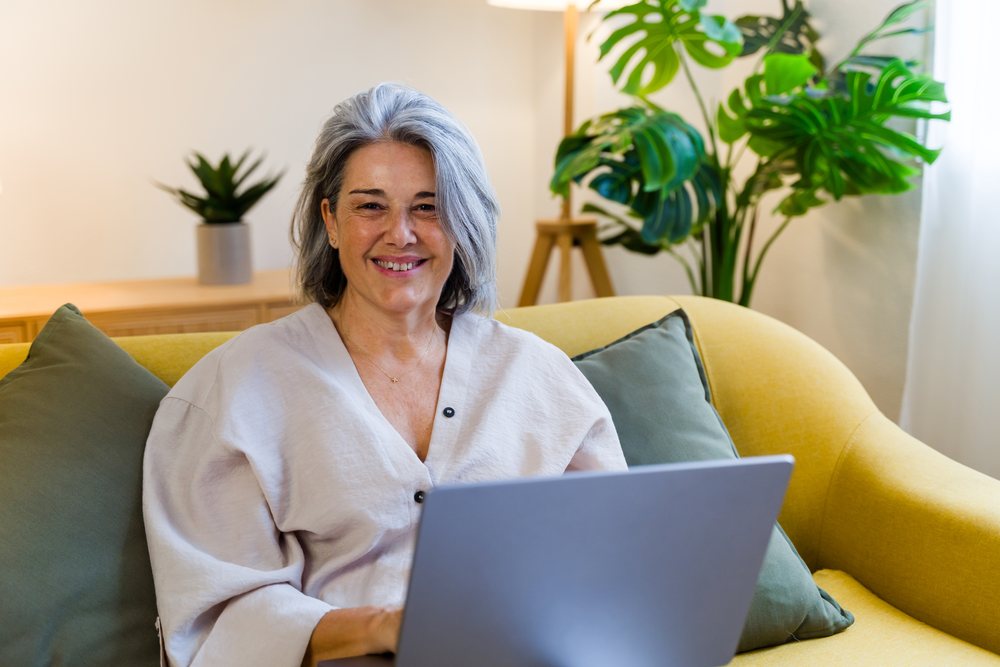 Middle-aged woman smiling while holding a laptop on a yellow couch indoors with green cushions, an indoor plant, and a floor lamp nearby, Spain.