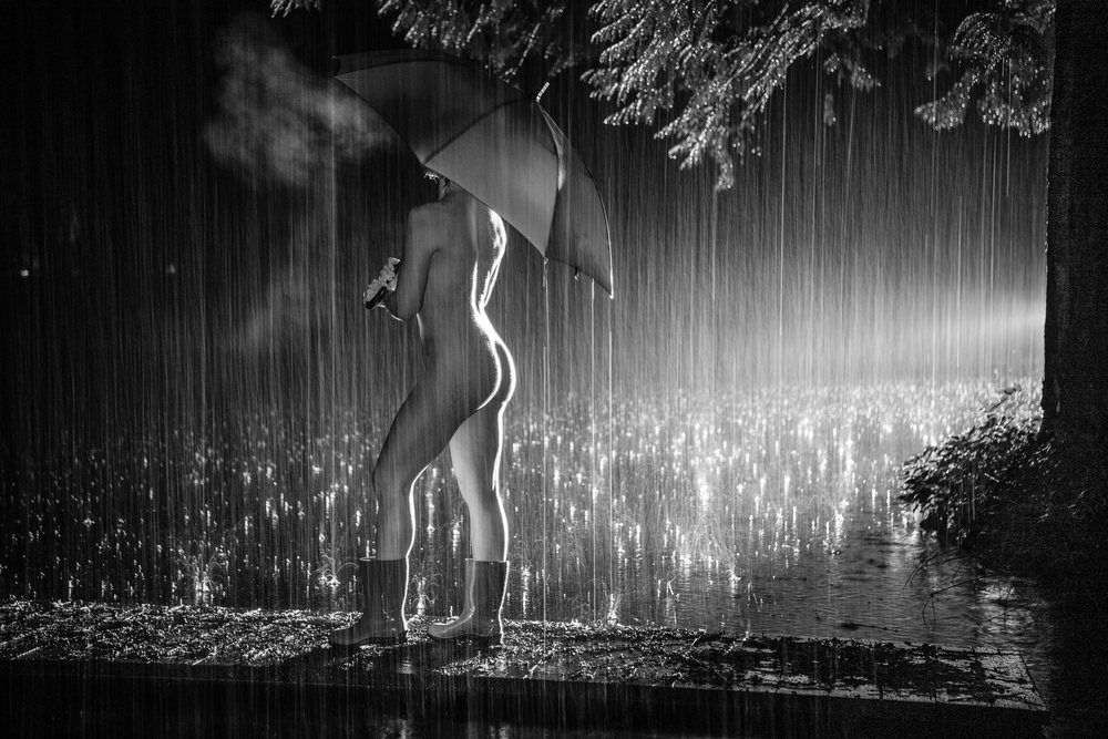 Nude woman stands outdoors at night in the rain holding an umbrella and wearing boots on wet ground with puddles, cosplay, nudity, freedom, expression, USA.