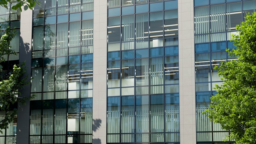 Office building with glass facade reflecting trees and nearby structures on a sunny day.