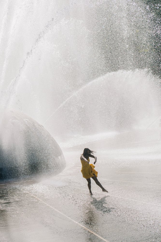Woman wearing a yellow dress is playing and dancing in an outdoor urban water fountain during daylight, USA.