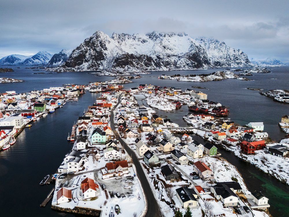 Aerial view showing snow-covered houses, boats docked in the harbor, and a mountainous landscape surrounding Henningsvaer village, Lofoten, Norway.