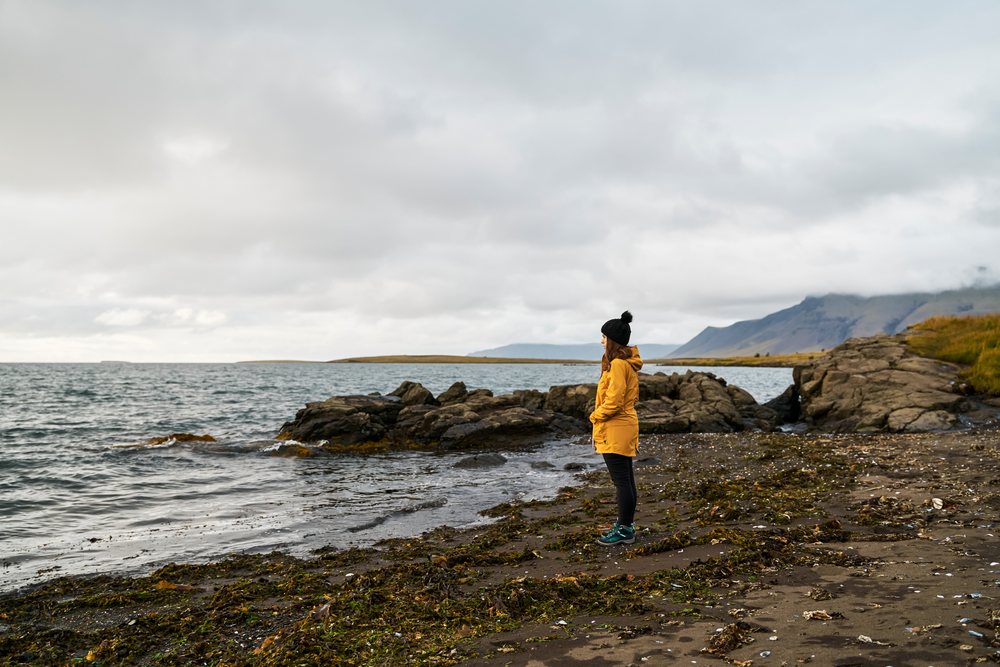A white woman in a yellow jacket stands on a rocky shoreline looking
