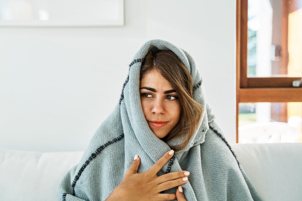 A young white woman wrapped in a cozy blanket indoors near a window