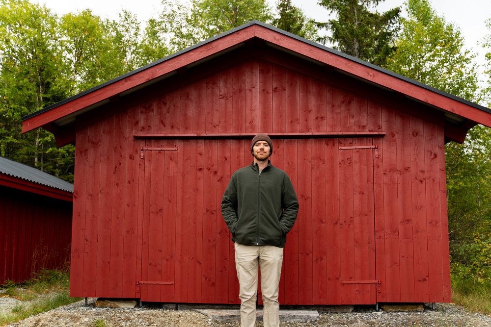 Man wearing green jacket and brown beanie stands in front of a red wooden cabin, Norway.