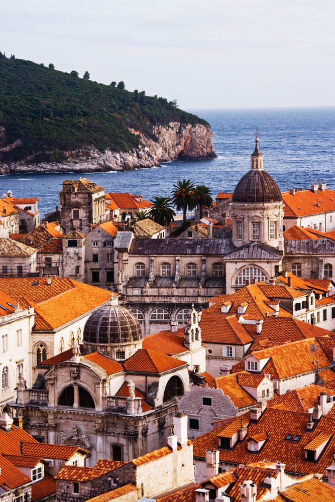 Scenic view of historic stone buildings with red tile roofs and a large domed church by the sea near Lokrum Island, Croatia.