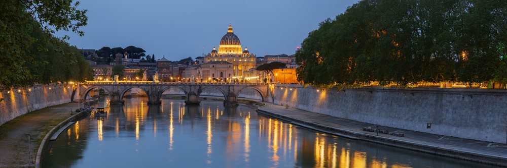 View of the Basilica of St Peter's at dusk across the Tiber River, with city lights reflecting on the water, Rome, Lazio, Italy.