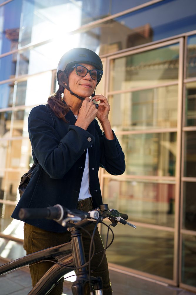 Woman putting on a helmet next to her bicycle on an urban sidewalk with reflective windows and sunlight, Málaga.