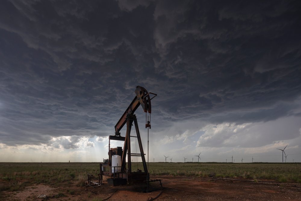 Pump jack operates under dark storm clouds with wind turbines visible on the Great Plains, USA, Tornado Alley.