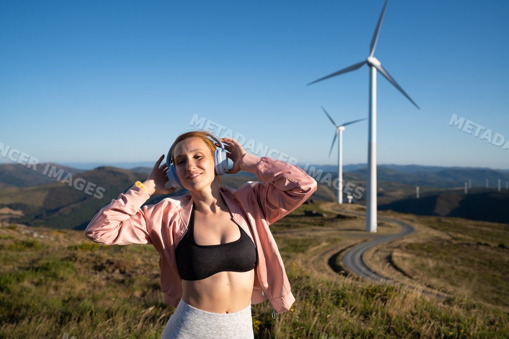 A white woman listens to music outdoors near wind turbines in Galicia