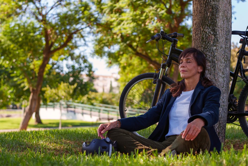 Woman peacefully meditating on grass beneath a tree in a sunny park with her bicycle and helmet nearby, Málaga.