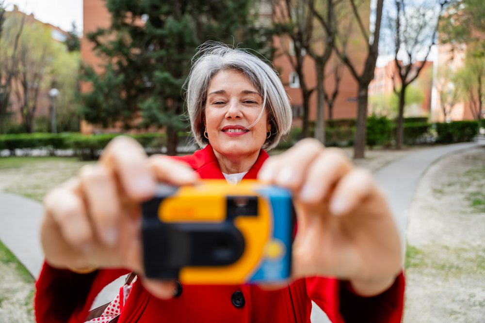 Woman with gray hair in a red coat smiles while holding a disposable camera in an urban park with trees and pathways, Spain.