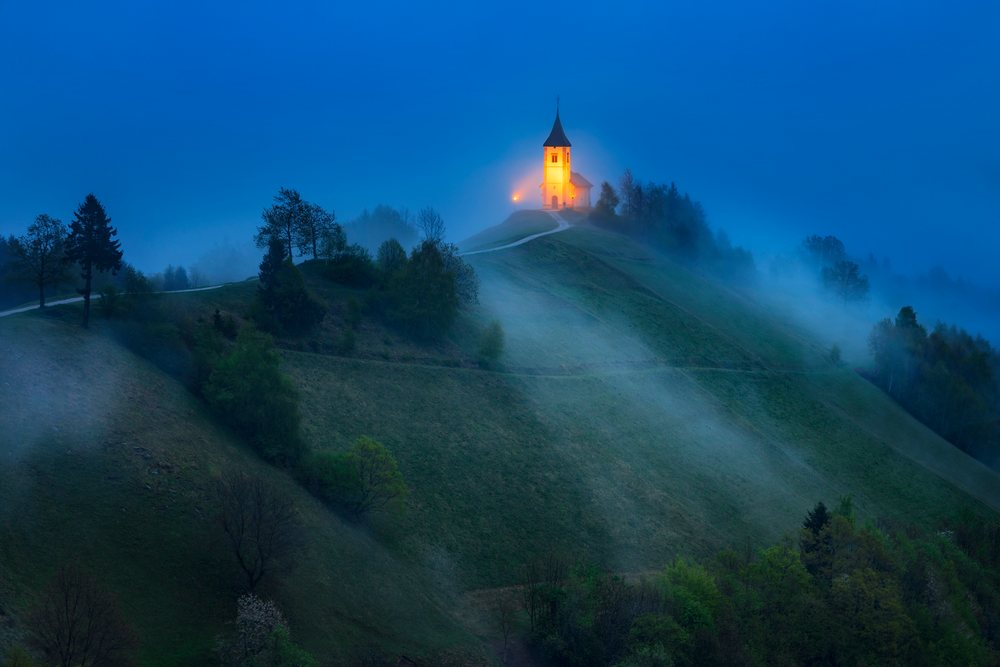 Illuminated Church of Primus and Felician stands on a hilltop with mist and trees at blue hour on Jelovica Plateau, Landscape, Jamnik, Slovenia.
