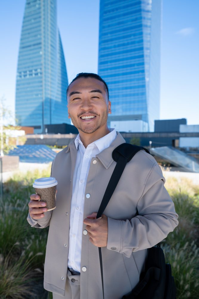 Man holding coffee cup and smiling outdoors in an urban area of Madrid with modern buildings in daylight, Spain.