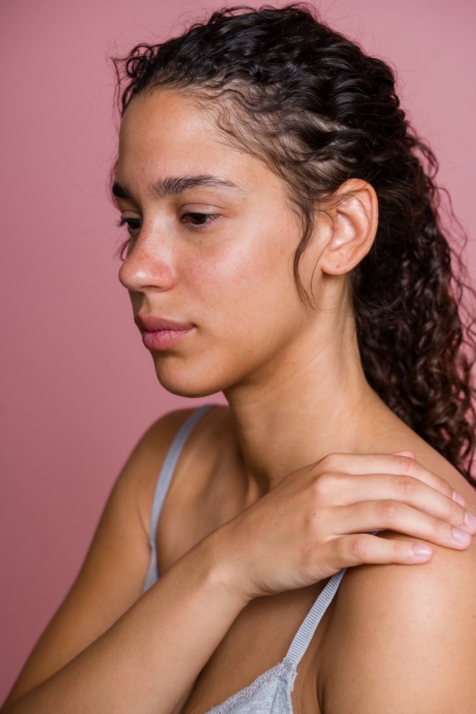 Woman with curly dark hair in profile resting her hand on her shoulder against a pink background portrait, beauty shoot, Spain.