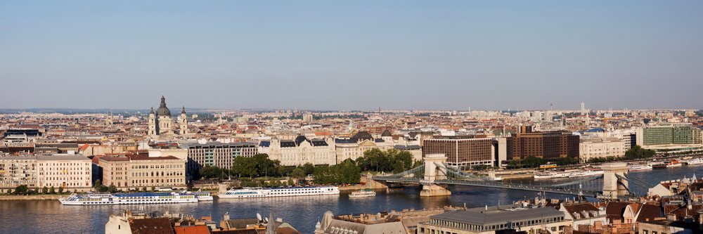 Cityscape of Budapest showing the Danube River with chain bridge, historic buildings, church dome, and river boats under a clear summer sky.