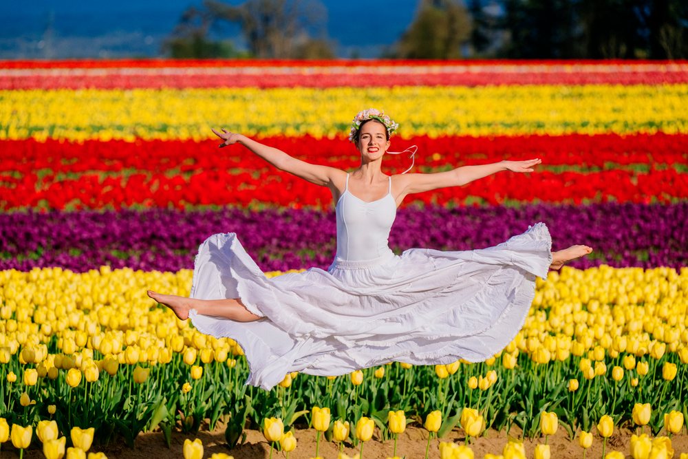 Ballet dancer jumps with arms outstretched in a vibrant tulip field in the USA wearing a white dress and flower crown on a clear spring day.
