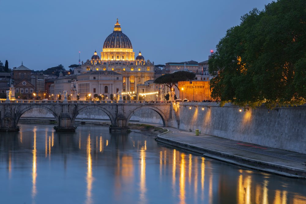Basilica of St Peter's illuminated at Dusk beside a river with a stone arched bridge and historic buildings, Rome, Lazio, Italy.
