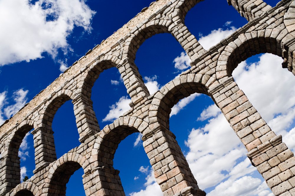 Roman aqueduct with multiple stone arches standing as an ancient historical monument, Spain under a partly cloudy blue sky, Segovia.