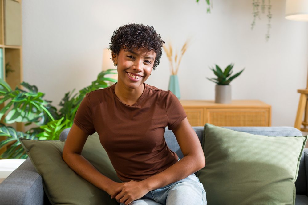 Young woman seated on a gray couch smiling indoors in a living room with green cushions and indoor plants.