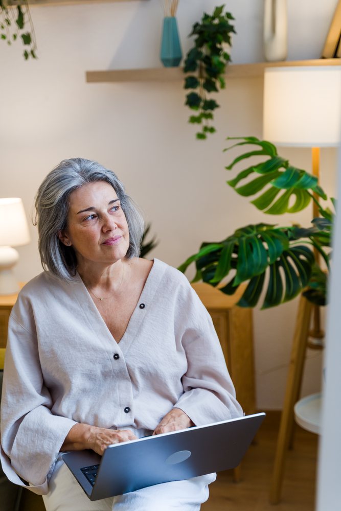 Woman with gray hair sits indoors using a laptop in a modern room with plants and lamps, Spain.