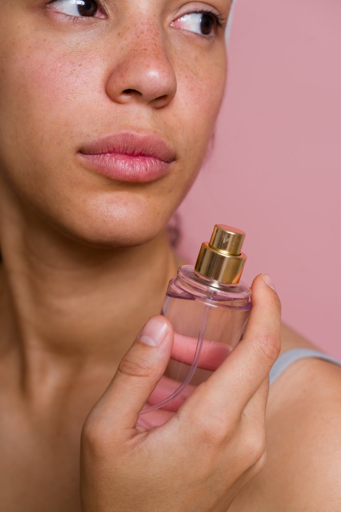 Woman with freckles holding a clear perfume spray bottle with gold nozzle against a pink background in a beauty shoot, Spain.