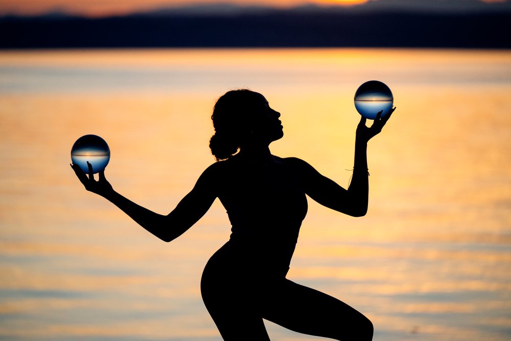 Silhouette of a woman holding two crystal balls balanced on her hands at sunset over water, yoga, acrobatics, USA.