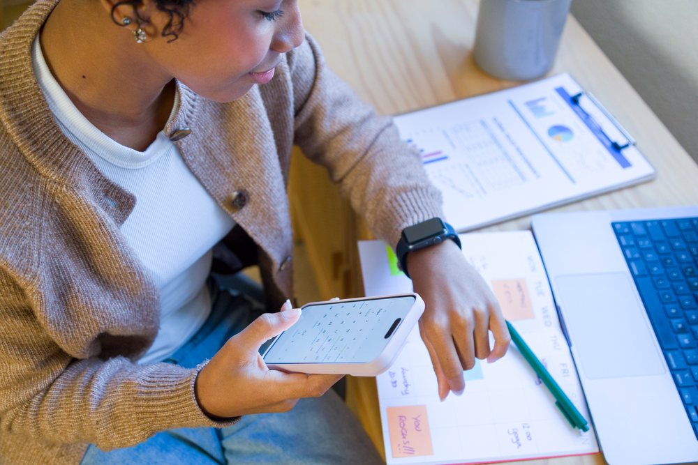 Woman holding smartphone with calculator app while working with notebook and laptop in home office, remote work.