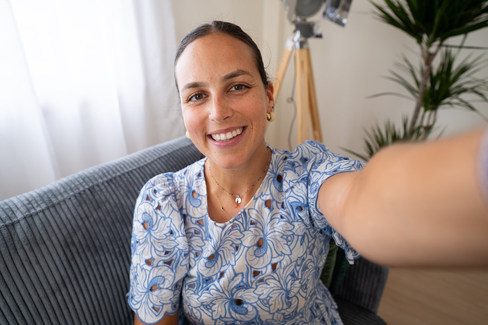 Woman smiling while taking a selfie on a gray upholstered couch indoors with a floor lamp and green plant nearby, telecommuting, Spain.
