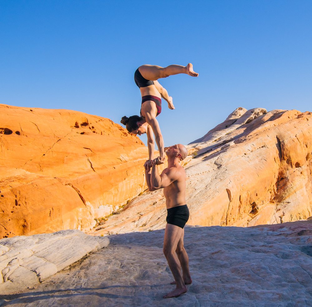 Man balancing a woman in a handstand yoga pose outdoors on rocky desert terrain under a clear blue sky, USA.
