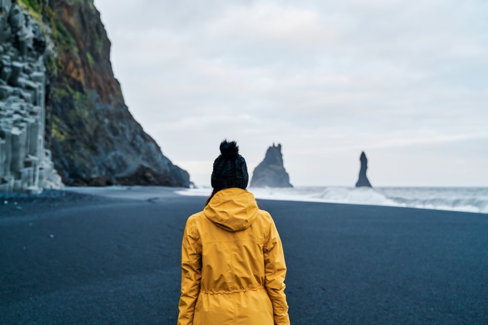 A white person in a yellow jacket walks on a black sand beach