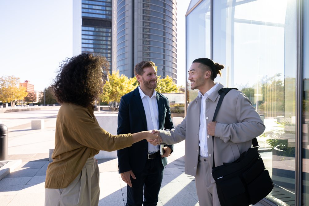 Three businesspeople—two men and one woman—smiling and shaking hands outdoors in front of a modern glass office building during daytime, Madrid, Spain.