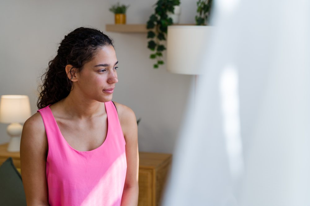 A woman in a pink tank top looks thoughtfully out of a window in a bright, modern room, Spain.