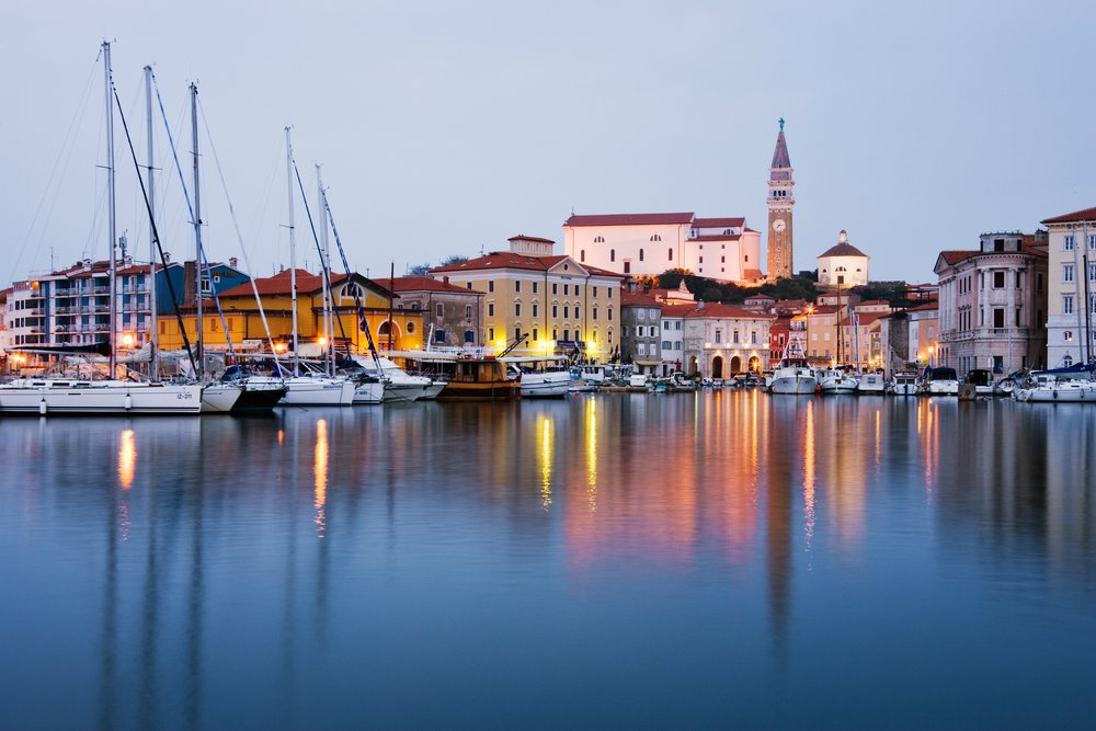 The Town of Piran waterfront shows sailboats docked along a marina with historic buildings reflecting on calm harbor waters, Dusk, Slovenia.