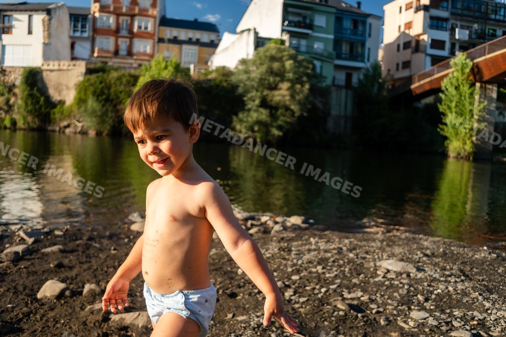 A toddler white boy plays by the riverbank with town buildings behind