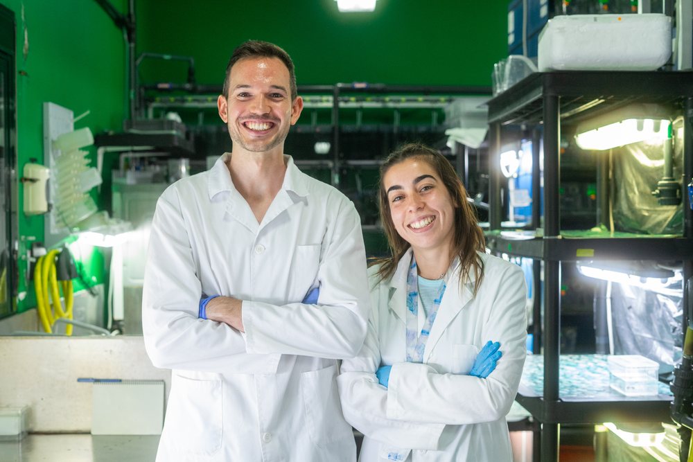 Two smiling scientists wearing white lab coats stand with crossed arms inside a laboratory research facility, Spain.