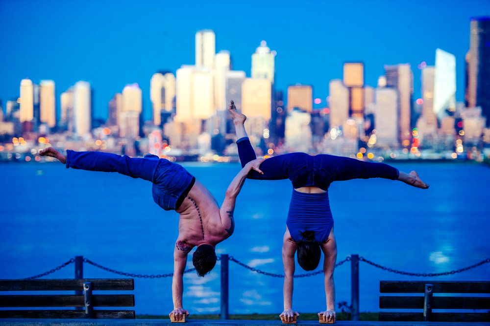 Two people performing partner acrobatics handstands by benches along the waterfront with the Seattle city skyline at evening, USA.