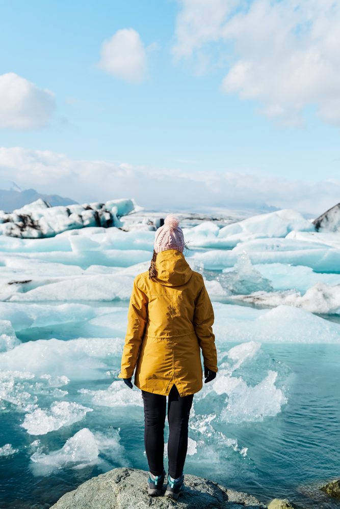 A woman wearing a yellow jacket and pink hat stands on a rock looking out over icy water and icebergs in a frozen landscape, Iceland.