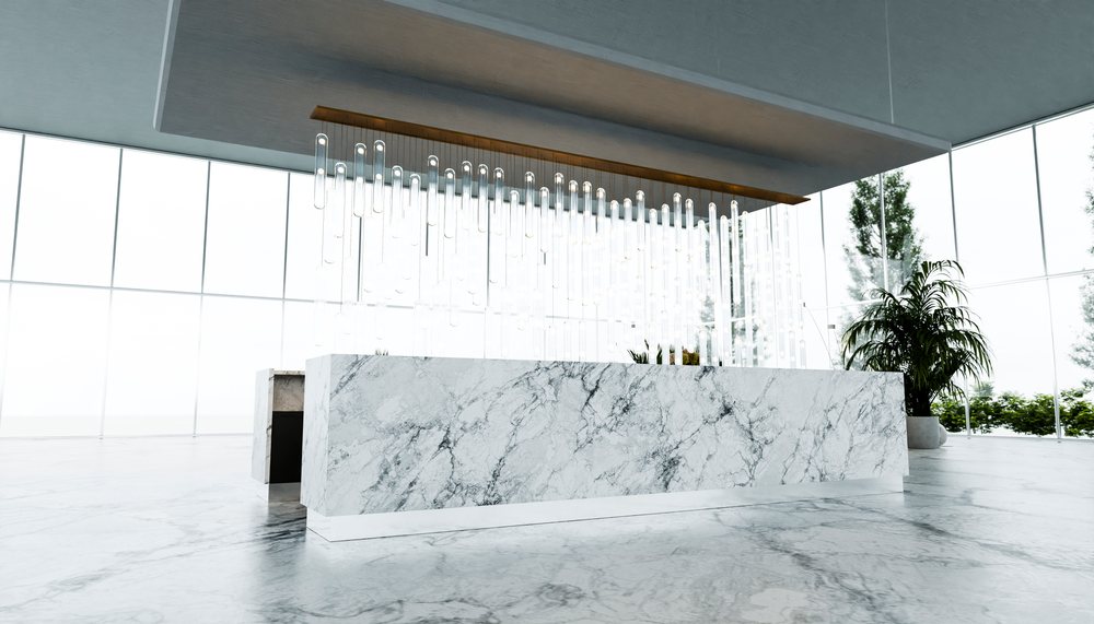 Spacious modern lobby with a large marble reception desk, hanging glass tubular lights, indoor plants, and floor-to-ceiling windows providing natural light.