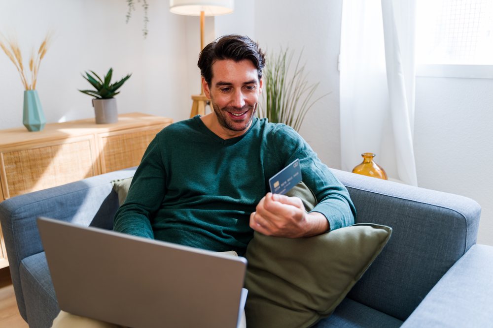 Man sitting on grey sofa with green pillow using laptop and holding a credit card in a well-lit living room with indoor plants and floor lamp, Spain.