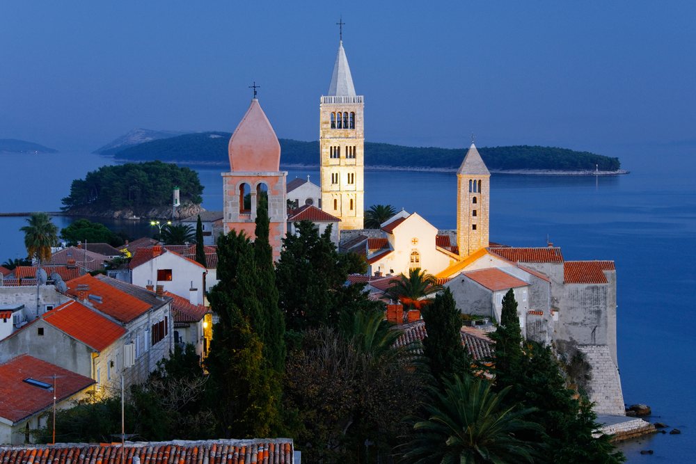 Historic church towers and red tiled roofs of Rab Island town with calm sea and islands in the evening light, Croatia.