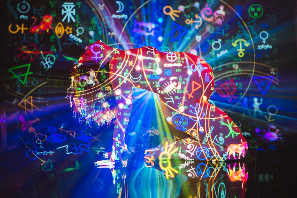 Female person kneeling with colorful astrology symbols projected on her body against a dark background with light rays and floor reflection, pregnancy.