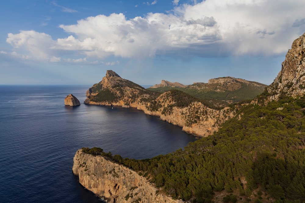 Rocky cliffs and green pine forest along Cap Formentor coastline overlooking the Mediterranean Sea under a partly cloudy blue sky, Landscape, Lifestyle, Pollença, Mallorca, Spain.